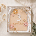 "Kiddospot wooden milestone card in a gift box, decorated with small wooden flowers and tied with twine, shown in a styled flat-lay setting."