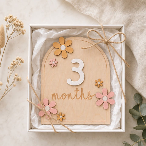 Kiddospot wooden milestone card in a gift box, decorated with small wooden flowers and tied with twine, shown in a styled flat-lay setting.