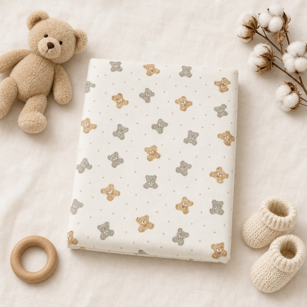A folded Kiddospot's fitted sheet with a repeating teddy bear print in beige and gray on an off-white background, styled in a flat lay with a plush teddy bear, cotton bolls, knit booties, and a wooden ring on a soft cream fabric backdrop.