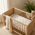 A light natural wood baby crib with vertical slats, styled in a bright nursery with a cream fitted sheet featuring a small bear pattern, a cream muslin throw blanket, and a plush teddy bear resting on the side.