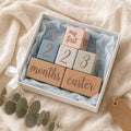 "A gift box containing Kiddospot's pastel wooden blocks with engraved text and numbers, including 'my first', '2', '3', 'months', and 'easter', arranged on light blue tissue paper. The box sits on a beige fabric surface with a white ribbon and eucalyptus sprig nearby."
