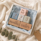 A gift box containing Kiddospot's pastel wooden blocks with engraved text and numbers, including 'my first', '2', '3', 'months', and 'easter', arranged on light blue tissue paper. The box sits on a beige fabric surface with a white ribbon and eucalyptus sprig nearby.