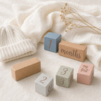 A set of Kiddospot's pastel wooden baby milestone blocks arranged on a soft cream fabric with a knitted cream hat and dried flowers. The blocks include labels for one year, months, two months, three months, three years, and my first birthday.