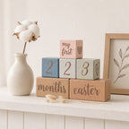 A set of Kiddospot's wooden baby milestone blocks stacked on a white shelf, including blocks that say '2', '2', '3', 'months', 'easter', and 'my first'. The blocks are in pastel blue, sage green, pink, and natural wood tones, styled with a ceramic vase of cotton stems and a framed botanical print.