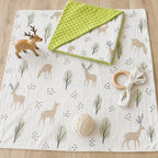 A Kiddospot baby blanket with a deer and tree pattern is laid out on a light wood floor, styled with a wooden toy deer, a folded green minky dot muslin cloth, a wooden ring rattle, and a knitted cream ball.