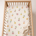 A Kiddospot fitted crib sheet with a cream-colored sheet featuring a delicate pastel floral pattern, shown inside a light wood crib with a folded cream textured blanket.