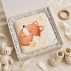 A folded baby blanket with a watercolor sleeping fox design in orange, cream, and white, presented in an open white gift box with ruffled tissue paper. The box is styled with a small teddy bear, wooden ring, and cream knit booties on a soft beige fabric background.