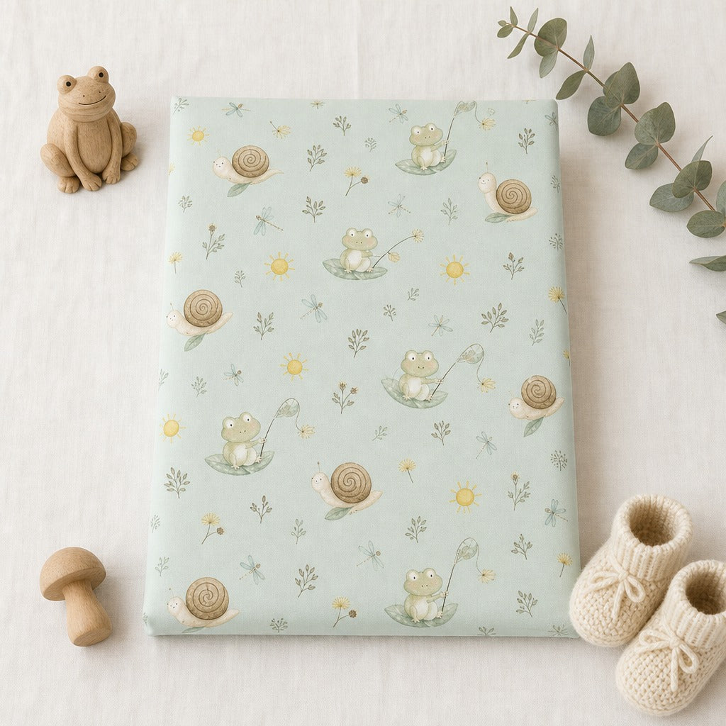 Kiddospot fitted sheet with a cute frog and snail pattern in soft sage green, cream, and yellow, styled in a flat-lay with a wooden frog, mushroom, baby booties, and eucalyptus leaves on a white fabric background.