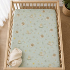 A Kiddospot fitted crib sheet with a light sage green fabric cover printed with a whimsical pattern of tadpoles, snails, and small flowers, shown inside a light wood crib with a folded cream blanket.