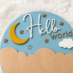 A round wooden wall plaque with a celestial night sky design, featuring a light blue background, a yellow crescent moon, brown stars, a white cloud, and the words 'Hello world' in white and brown. The bottom edge has a scalloped wooden border, and the plaque is displayed on a white faux fur surface.