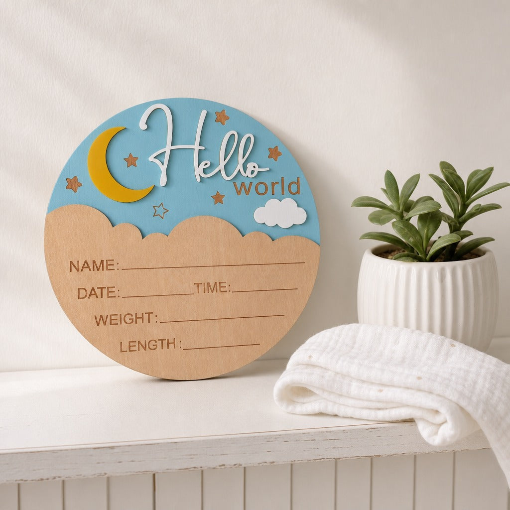 A round wooden baby milestone plaque with a blue sky and moon design, displayed on a white shelf next to a small potted plant and a folded white cloth.