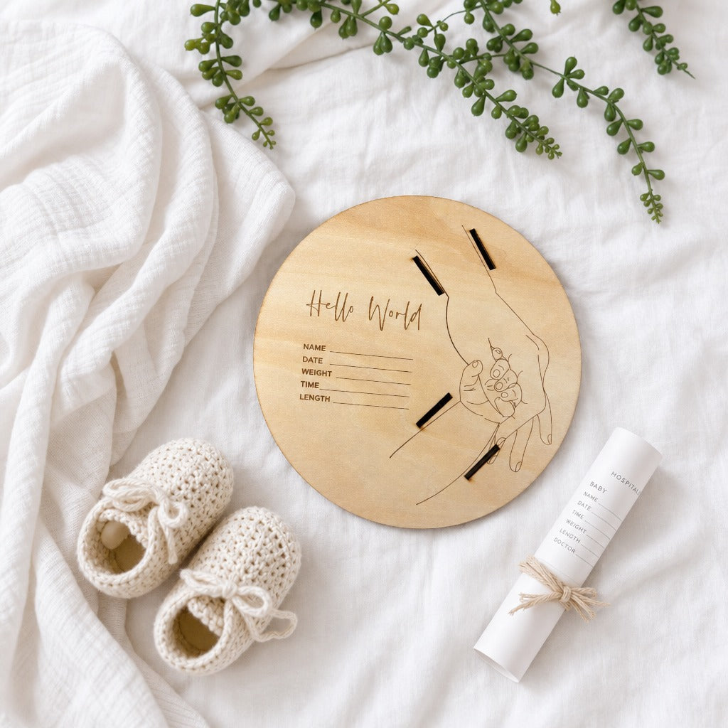 Kiddospot round wooden baby keepsake plaque engraved with 'Hello World' and baby details, styled on a white textured blanket with cream knit booties and a rolled white certificate tied with twine.
