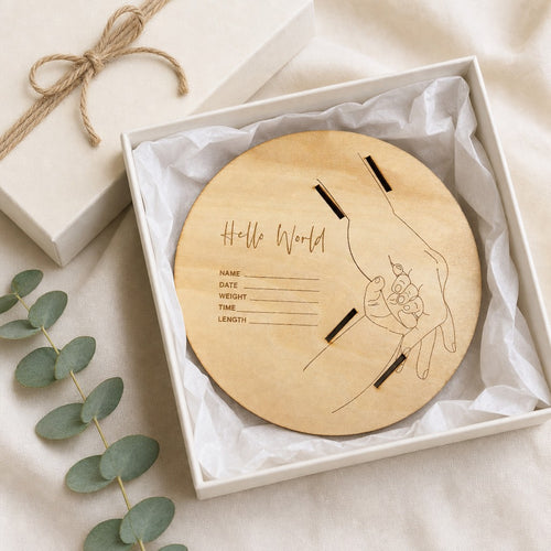 Kiddospot round wooden keepsake plaque engraved with 'Hello World' and a hand holding a flower, displayed inside a white gift box with tissue paper. A matching cream gift box with twine is visible in the background, along with eucalyptus leaves on a beige fabric surface.