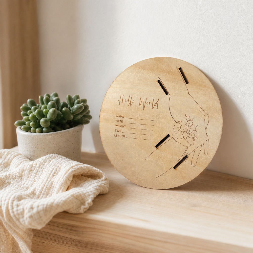 Kiddospot round wooden keepsake plaque engraved with 'Hello World' and a hand illustration, leaning on a light wood shelf next to a small potted succulent and a cream knit throw blanket.