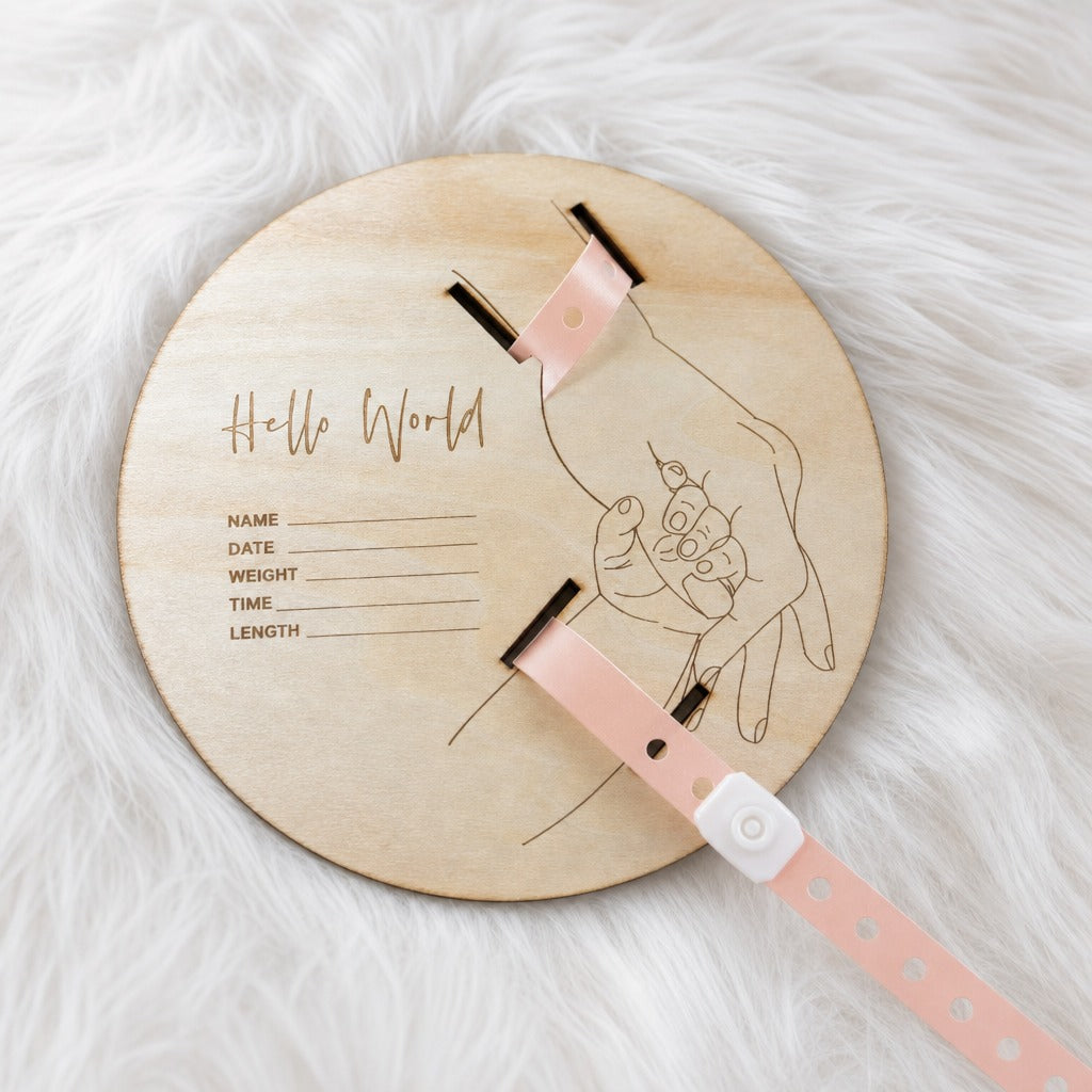 Kiddospot personalized wooden baby milestone plaque on a white faux fur background. The round, light-colored wooden plaque features an engraved line drawing of a baby in a cradle and the text 'Hello World' along with fields for name, date, weight, and time.