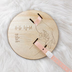 Kiddospot personalized wooden baby milestone plaque on a white faux fur background. The round, light-colored wooden plaque features an engraved line drawing of a baby in a cradle and the text 'Hello World' along with fields for name, date, weight, and time.