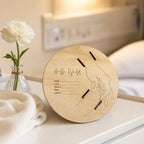 Kiddospot round wooden baby milestone plaque with engraved lines and text, styled on a bedside table next to a white flower in a glass vase and a folded cream knit blanket.