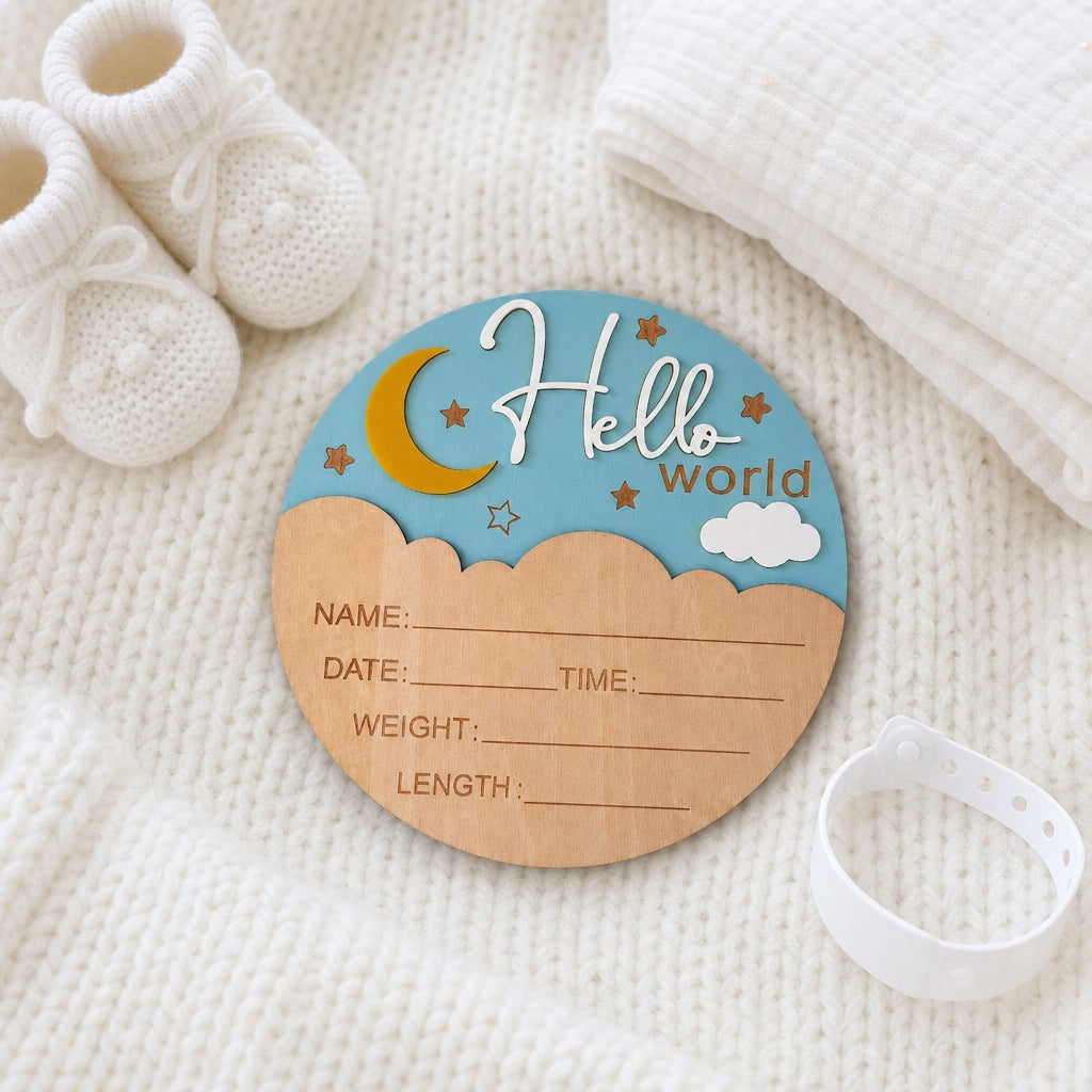 A personalized baby birth announcement plaque shaped like a moon and stars sky, placed on a white knit blanket with baby booties, a muslin cloth, and a white silicone band nearby.