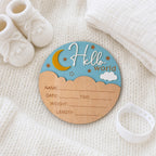 A personalized baby birth announcement plaque shaped like a moon and stars sky, placed on a white knit blanket with baby booties, a muslin cloth, and a white silicone band nearby.