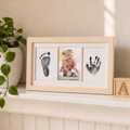 "Inkless baby handprint and footprint kit in natural wood frame with white mat insert, displayed on a nursery shelf with soft decor"