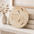 Kiddospot wooden baby announcement sign bracelet holder with engraved text on a white surface with a vase of cotton flowers.