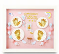 Decorative shadow box with gold hand and footprints, colorful beads, and baby-themed elements on a pink background.