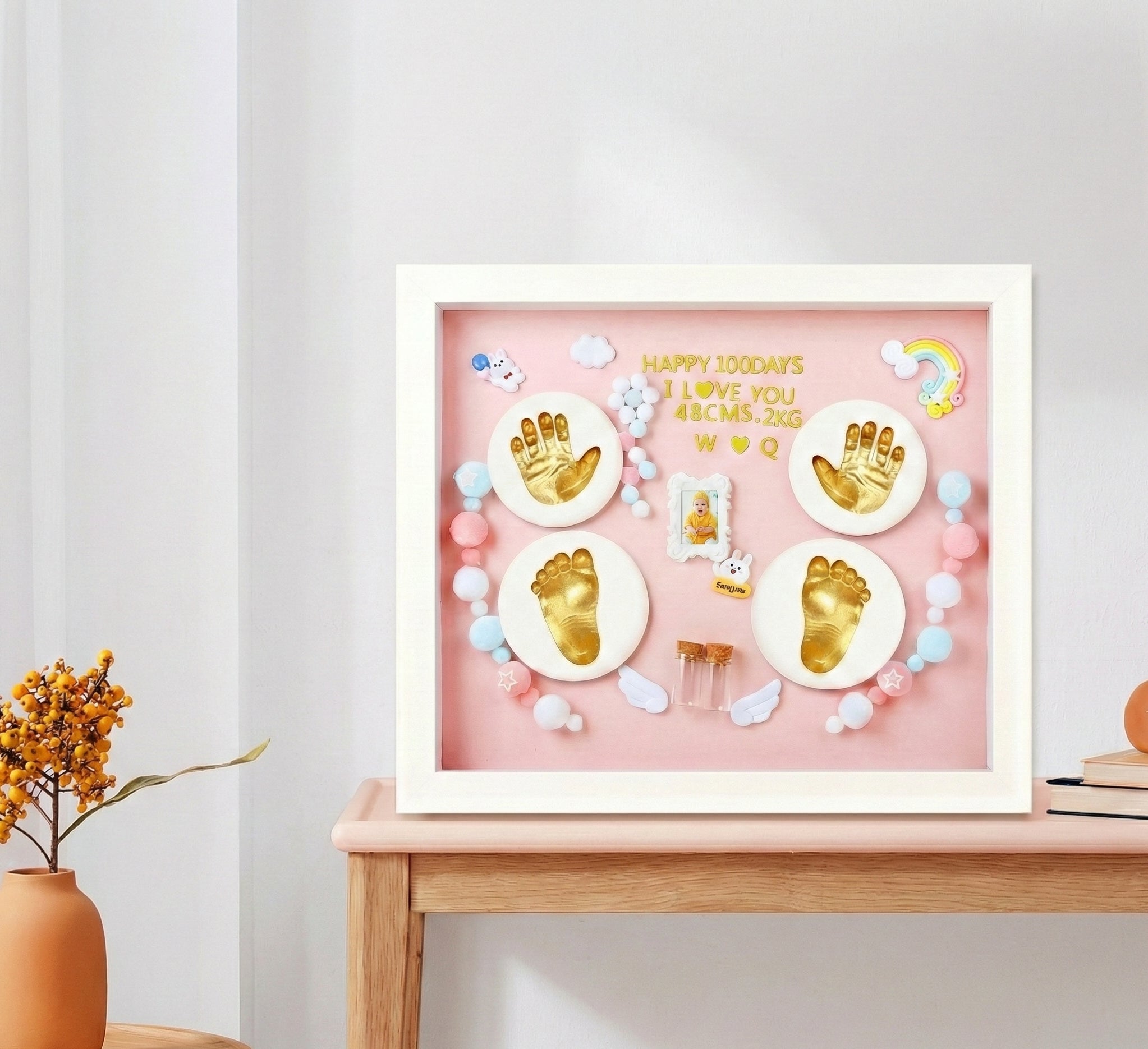 Decorative shadow box with gold hand and footprints on a pink background, placed on a wooden surface.
