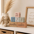 Kiddospot baby milestone blocks with numbers and text on a shelf with a plush bunny and framed picture.