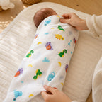 Parent's hands swaddling a newborn baby with the Kiddospot GOTS certified organic muslin swaddle in dinosaur print — white double-gauze cotton with colourful cartoon dinosaurs in kelly green, sky blue, yellow, orange and purple. Soft warm nursery setting.