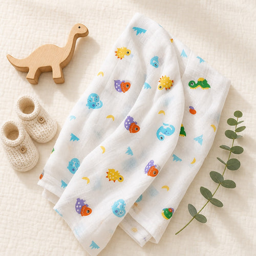 Overhead flat lay of the Kiddospot GOTS organic cotton muslin swaddle in dinosaur print — white double-gauze fabric with colourful cartoon dinosaurs styled with a wooden dinosaur toy, cream knitted baby booties and eucalyptus. Perfect gender-neutral baby shower gift.