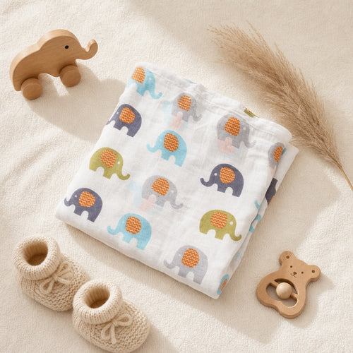 Overhead flat lay of the Kiddospot GOTS organic cotton muslin swaddle in elephant print — white double-gauze fabric with colourful elephant motifs styled with nursery props including a wooden toy and cream knitted booties. Perfect organic baby shower gift.