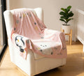 Pink baby blanket with cartoon designs draped over a chair in a room with a plant and crib.