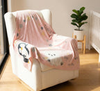 Pink baby blanket with cartoon designs draped over a chair in a room with a plant and crib.
