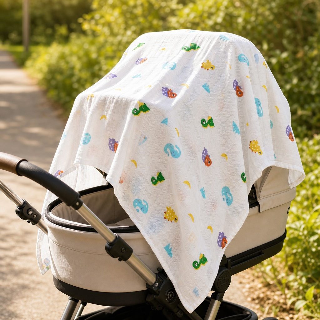 Kiddospot GOTS organic cotton muslin swaddle in dinosaur print draped over a pram as a sun shade — demonstrating the multi-use versatility of the 120x120cm breathable newborn wrap. Bright outdoor setting with natural light.