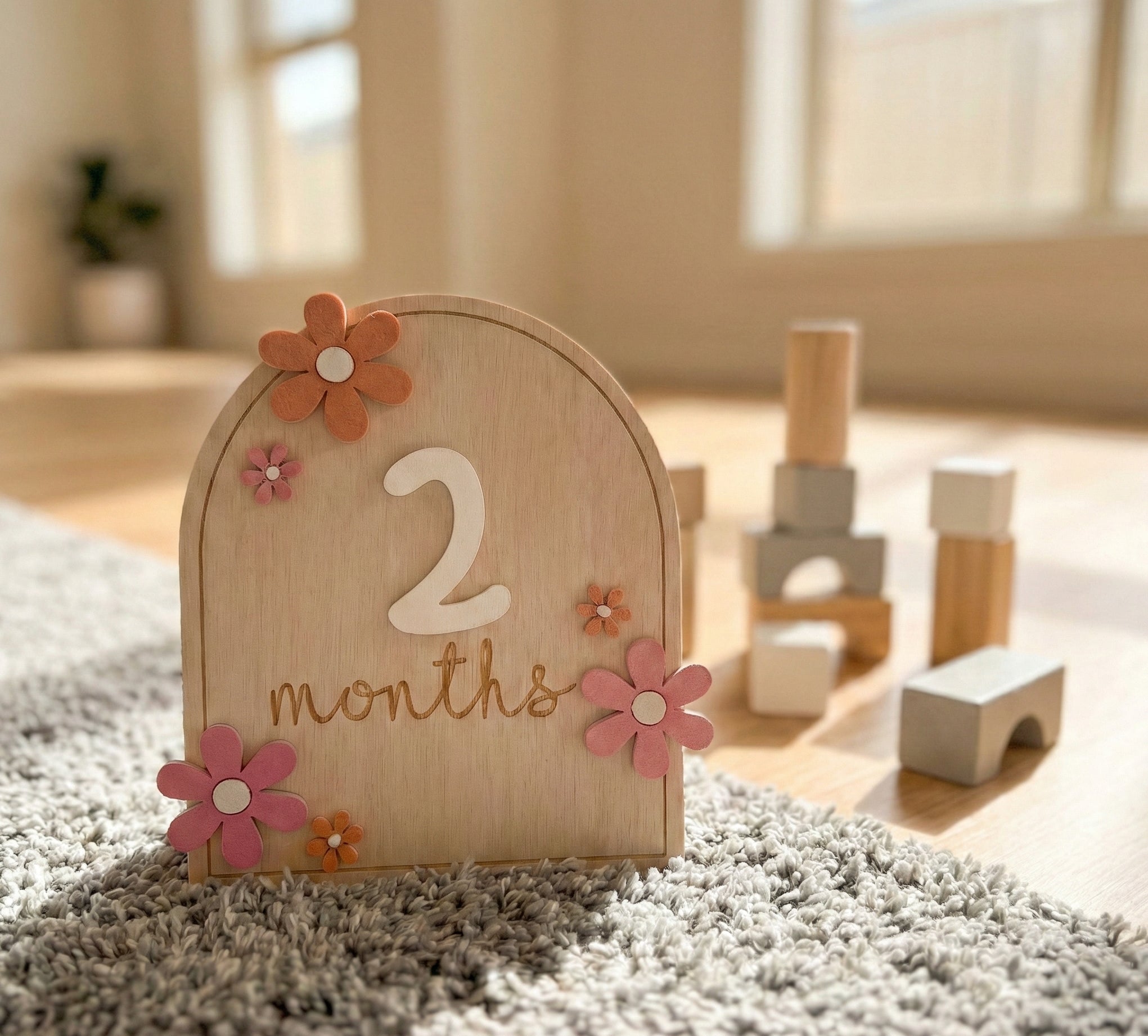 Wooden milestone with flowers and '2 months' text on a carpeted floor with toys in the background