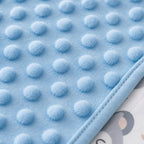Close-up of a Kiddospot light blue textured blanket with raised circular dots and a stitched border, partially layered over a printed baby fabric underneath.