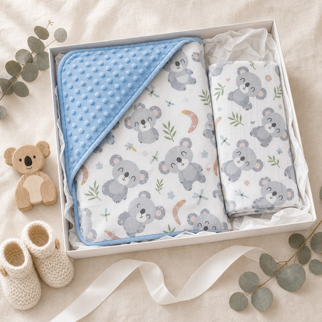 A baby gift set in a white box featuring a Kiddospot light blue minky dot blanket, a white muslin cloth with a gray koala print, and a small plush koala toy, styled with cream knit booties, eucalyptus leaves, and a ribbon on a beige fabric background.