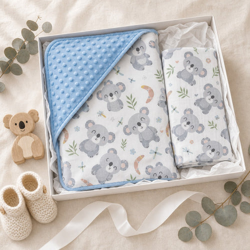A baby gift set in a white box featuring a Kiddospot light blue minky dot blanket, a white muslin cloth with a gray koala print, and a small plush koala toy, styled with cream knit booties, eucalyptus leaves, and a ribbon on a beige fabric background.