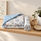 A folded and rolled Kiddospot muslin blanket with a koala print and blue dotted reverse side, styled on a wooden shelf next to a small teddy bear, a potted plant, and a framed print.