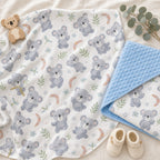 A Kiddospot baby blanket and swaddle with a repeating koala print, laid flat on a soft cream fabric background. One folded corner shows a light blue textured reverse side.