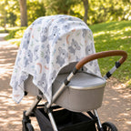 A baby stroller in a park setting, covered with a Kiddospot white muslin canopy print featuring gray koalas, blue flowers, and orange crescent moons. The stroller has a gray bassinet, silver frame, and a brown leather-look handlebar.