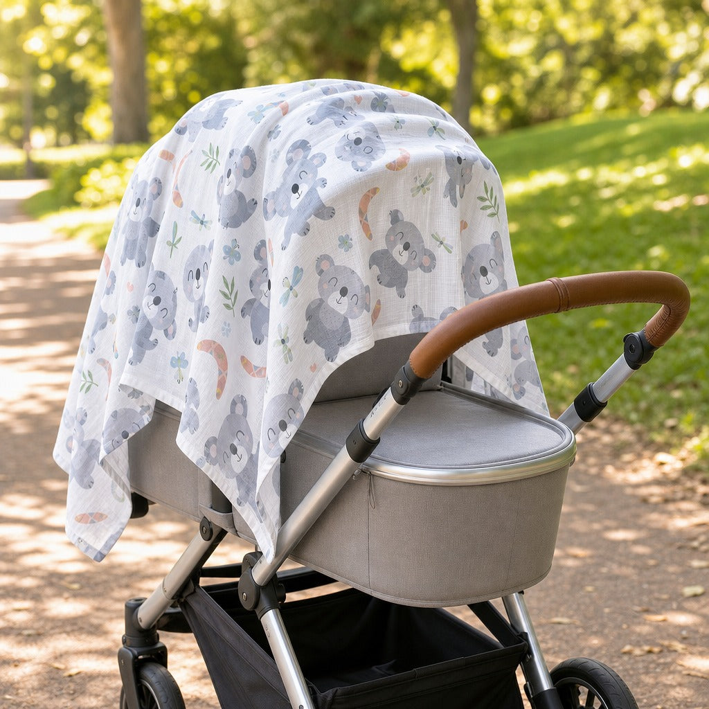 A baby stroller in a park setting, covered with a Kiddospot white muslin canopy print featuring gray koalas, blue flowers, and orange crescent moons. The stroller has a gray bassinet, silver frame, and a brown leather-look handlebar.