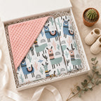 A baby gift set in a white box featuring a Kiddospot pink minky dot blanket and a matching patterned baby wrap with llamas and cacti, styled with a small cactus in a pot and knit baby booties on a beige fabric background.