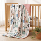 A Kiddospot baby blanket with a colorful llama and cactus pattern draped over a light wood crib, featuring a pink minky dot reverse side, in a bright nursery setting.