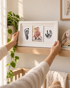 Parent hands placing a white triple-window inkless newborn handprint and footprint keepsake frame on a nursery shelf with plant and soft morning light