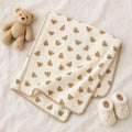 "A cream-colored baby blanket with a repeating pattern of small brown teddy bear faces, styled on a white textured fabric background alongside a plush teddy bear and a pair of cream knit booties."
