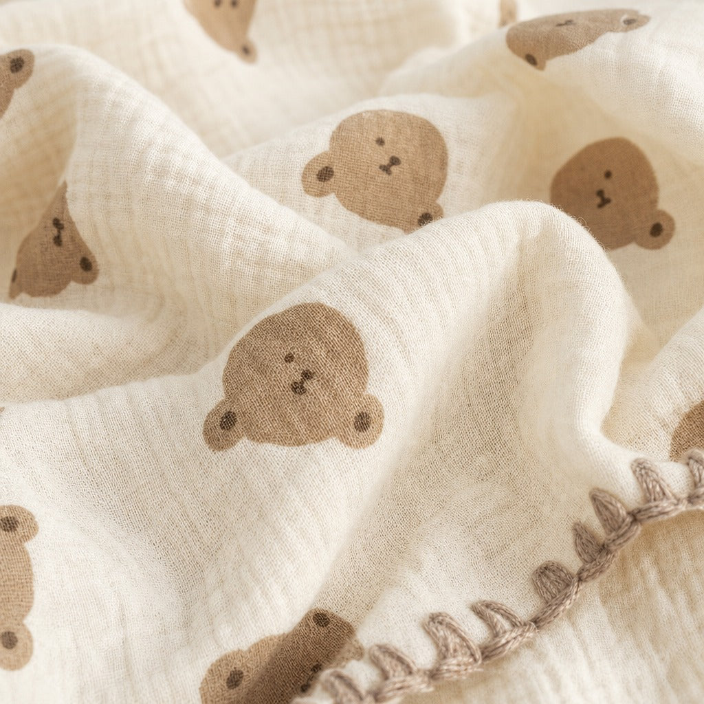 Close-up of a soft cream-colored muslin fabric with a repeating pattern of small brown teddy bear faces and a matching brown embroidered border.