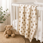 A cream-colored baby blanket with a repeating brown teddy bear pattern is draped over a white wooden crib in a softly lit nursery. A plush teddy bear and a potted eucalyptus plant sit on a woven rug nearby.