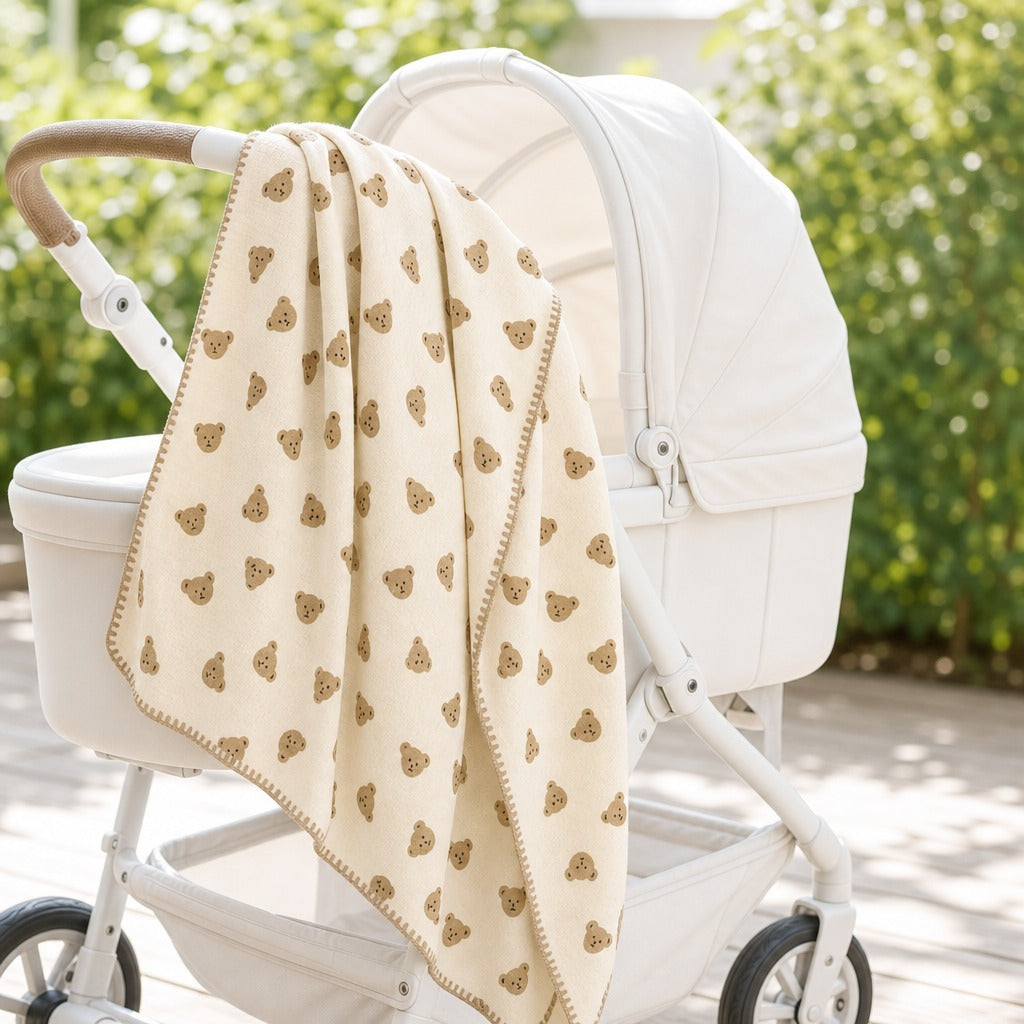 A cream-colored baby blanket with a pattern of small brown teddy bear faces and a matching brown border, draped over the handle of a white stroller in an outdoor garden setting.