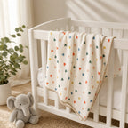 A cream-colored muslin baby blanket with a colorful heart pattern and a beige tassel border, draped over a white wooden baby crib. The scene is styled with a gray plush elephant toy and a potted eucalyptus plant in a bright, softly lit nursery.